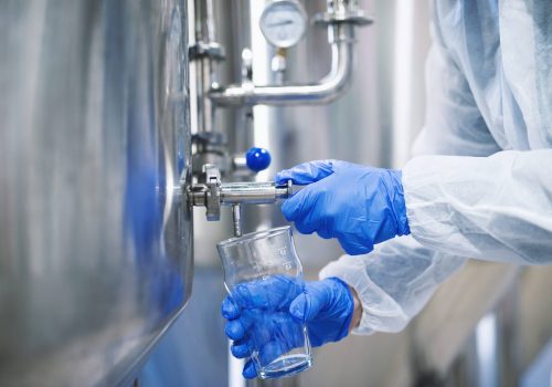 Close up view of hands in protective gloves opening tap valve and filling glass with samples from industrial machine reservoir.